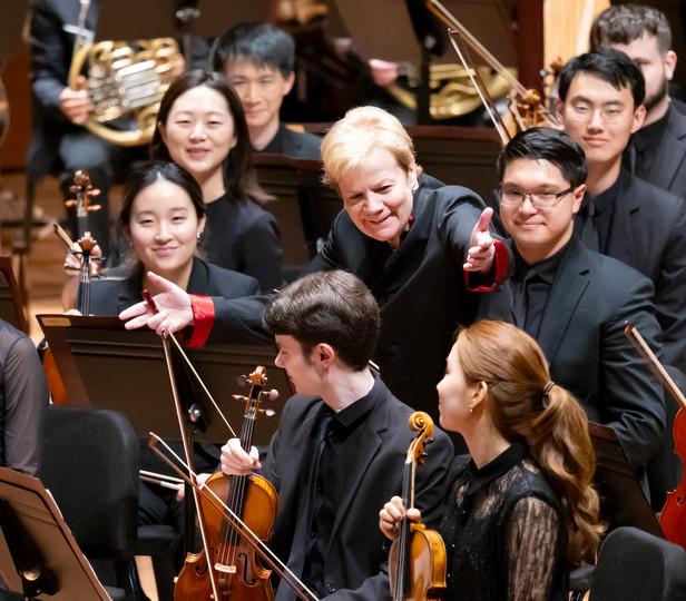 Alsop is engaging with the musicians of an orchestra, who are seated 和 holding their instruments, with smiles 和 attentive expressions on their faces.